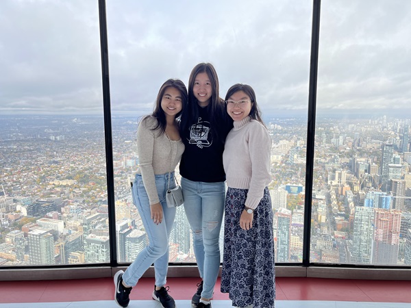 Three women stand in front of a floor to ceiling glass window over looking a city scape